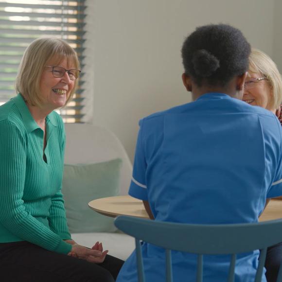 Woman in green smiles, talking with a nurse and another woman at a table.