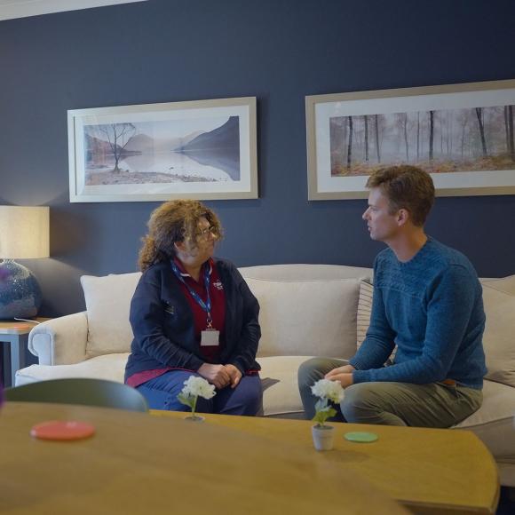 Woman in uniform talks to a man on a white sofa in a modern living room.