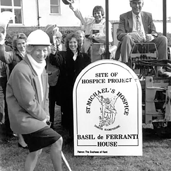 Woman in hard hat digging at groundbreaking for St Michael's Hospice, group looking on.