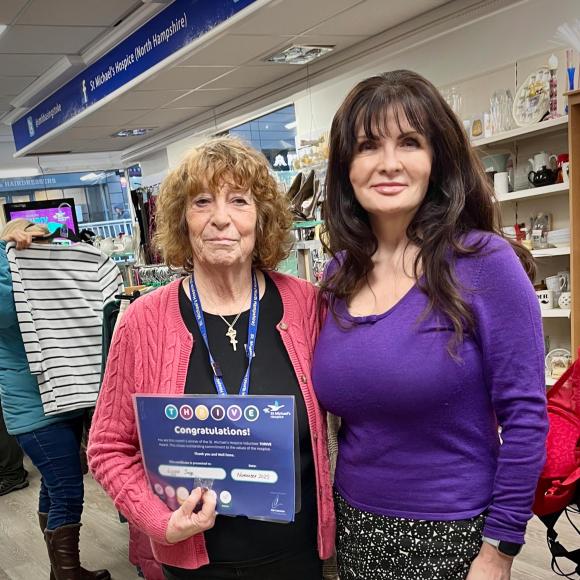 Older woman in pink holds a certificate next to a younger woman in purple, inside a shop.