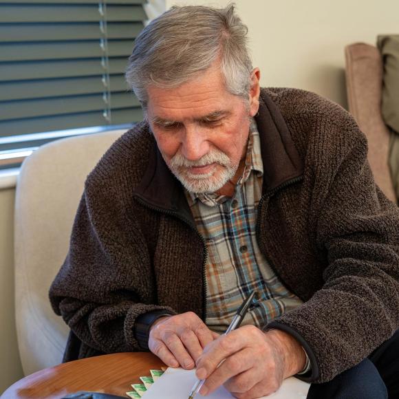 Grey-haired man in a brown jacket writing intently.
