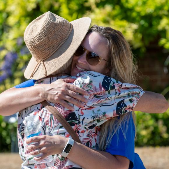 Two people sharing a warm hug outdoors, one in a hat, the other in sunglasses.