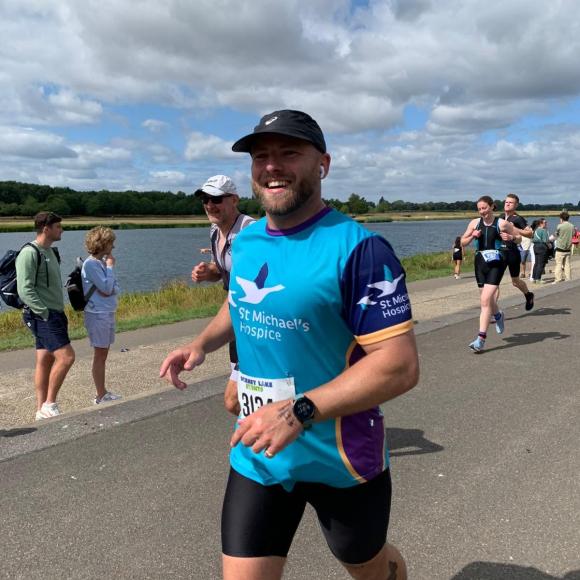 Smiling man in blue hospice shirt running a race by a river.