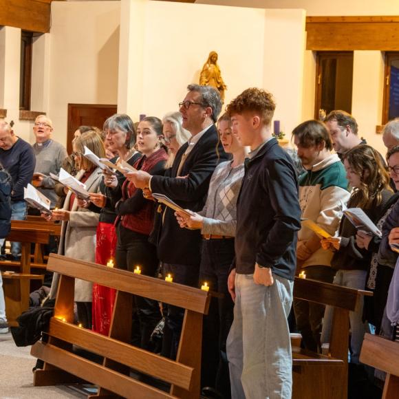 Congregation standing and singing from papers in a church, some holding lit candles.