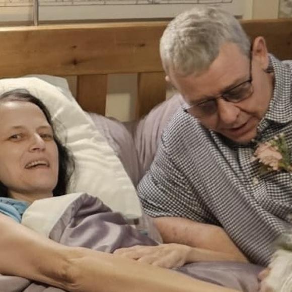 Man with a boutonnière leans over a smiling woman in bed.