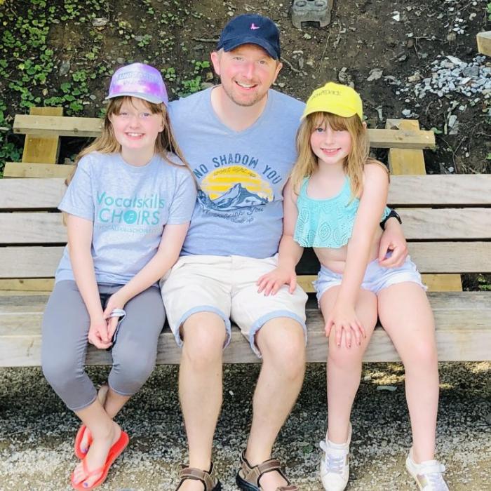 Smiling man and two young girls in hats sitting on a wooden bench outdoors.
