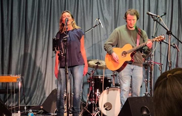 Two musicians perform on stage; a woman sings into a microphone, a man plays acoustic guitar.