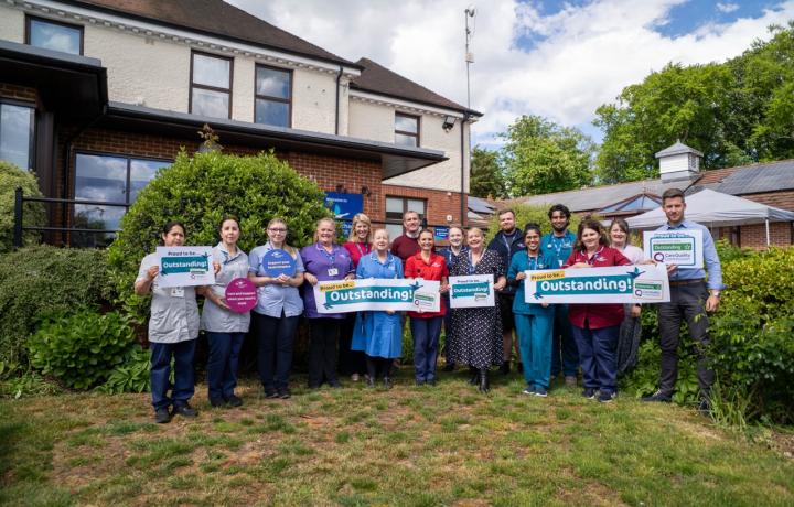 Group of smiling healthcare staff holding "Outstanding" signs in front of a care facility.