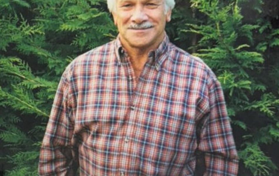 Smiling man with white hair and mustache, wearing a plaid shirt, green background.