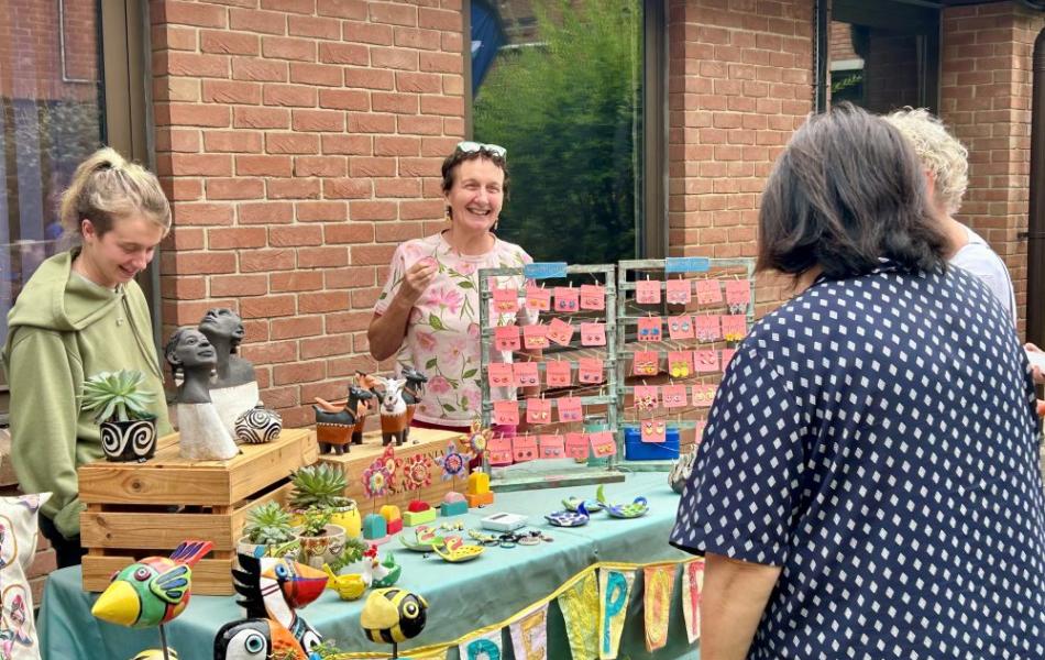 Woman smiles behind a colorful craft stall with handmade birds and plants.