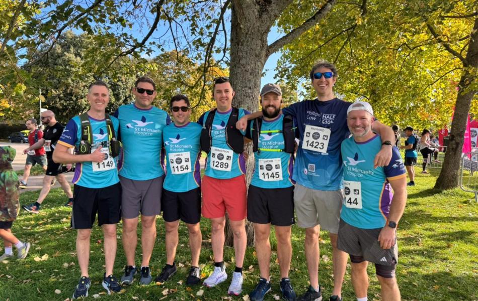 Seven smiling men in blue race shirts with bibs pose under a tree at a running event.
