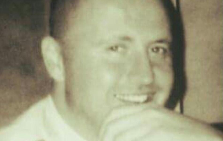 Sepia photograph: Smiling man with shaved head, hand to chin.