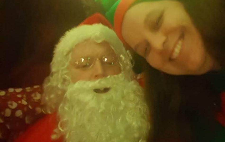 Santa Claus and a smiling woman in an elf hat under colorful festive lights.