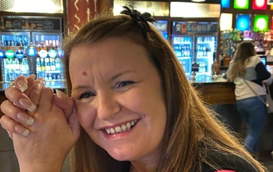 Smiling woman with long hair and clasped hands in a pub, bottles and taps in background.