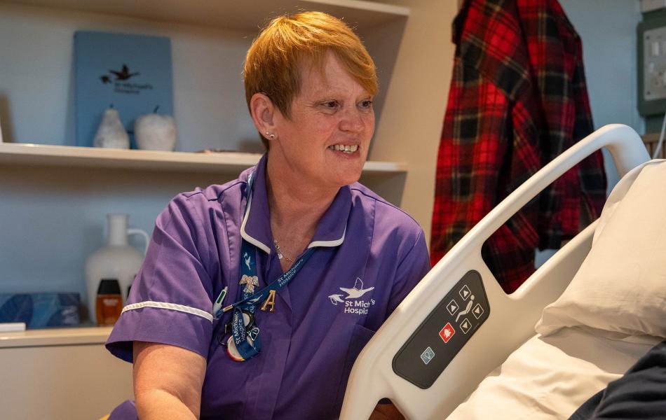Friendly nurse in purple uniform talks to a patient in a hospital bed.