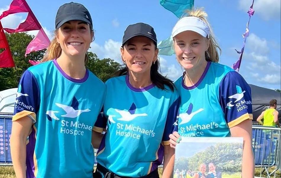 Three smiling women in St Michael's Hospice shirts; one holds a photo, outdoors.