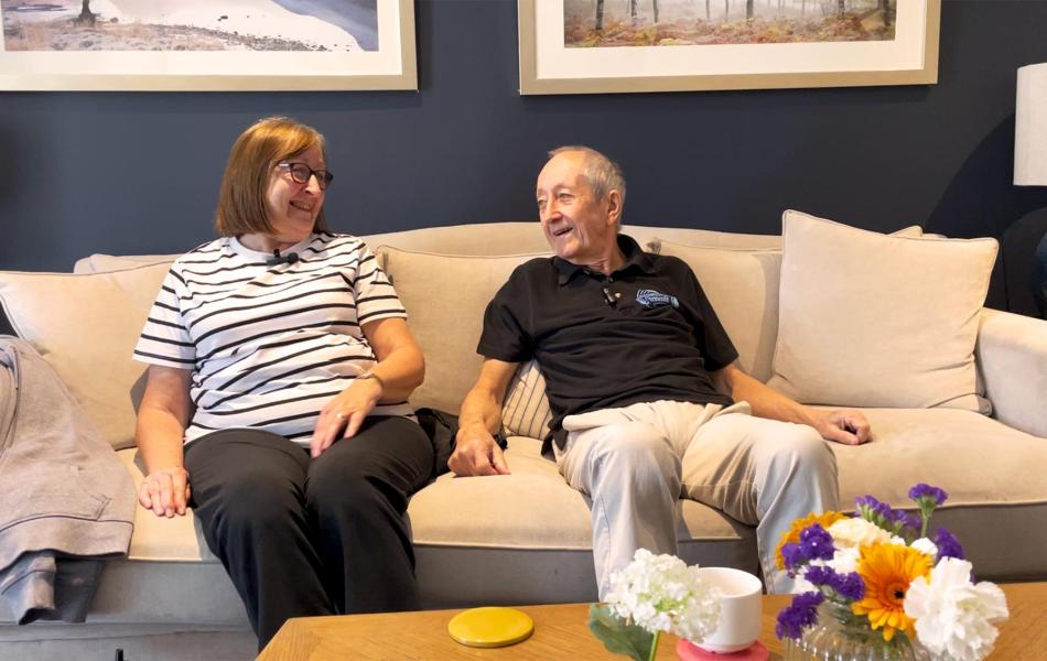 Elderly couple happily talking on a couch in a living room with flowers.