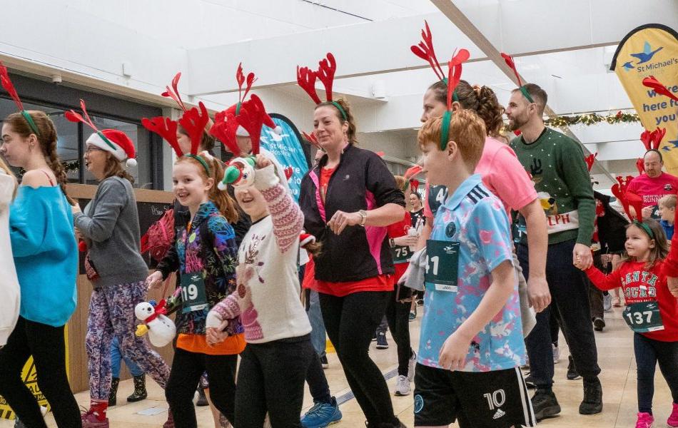 Adults and children in Christmas outfits, some with reindeer antlers, at a festive indoor event.