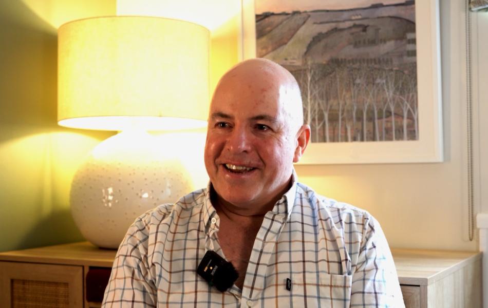 Man in plaid shirt smiles, wearing a lapel mic. Warm room with lamp and artwork.