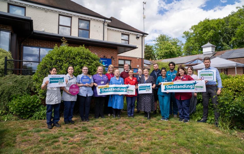 Group of smiling healthcare staff holding "Outstanding" signs in front of a care facility.