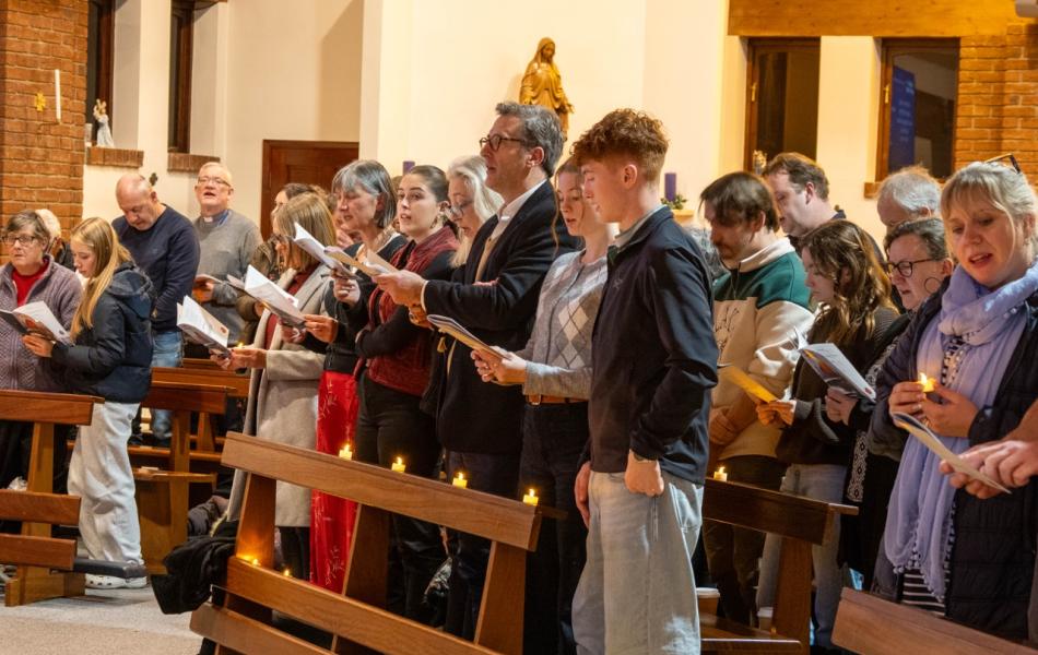 Congregation standing and singing from papers in a church, some holding lit candles.