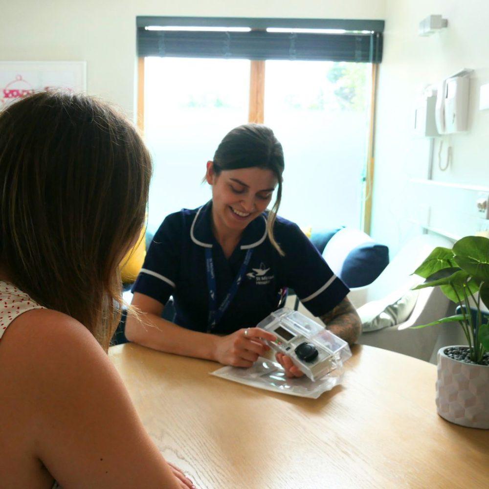 Smiling nurse in uniform shows a packaged medical device to a patient in a clinic room.