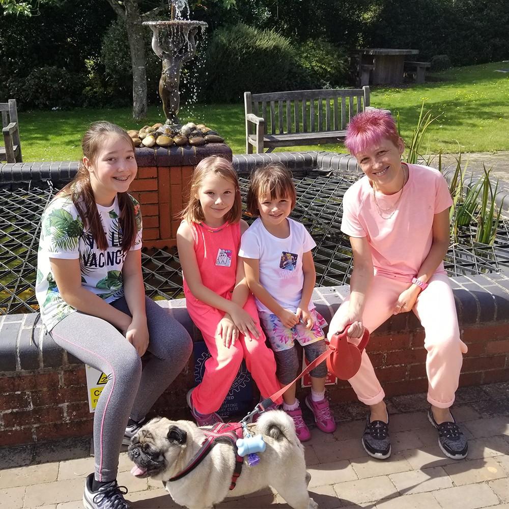 Adult with pink hair, three girls, and a pug sit by a park fountain.