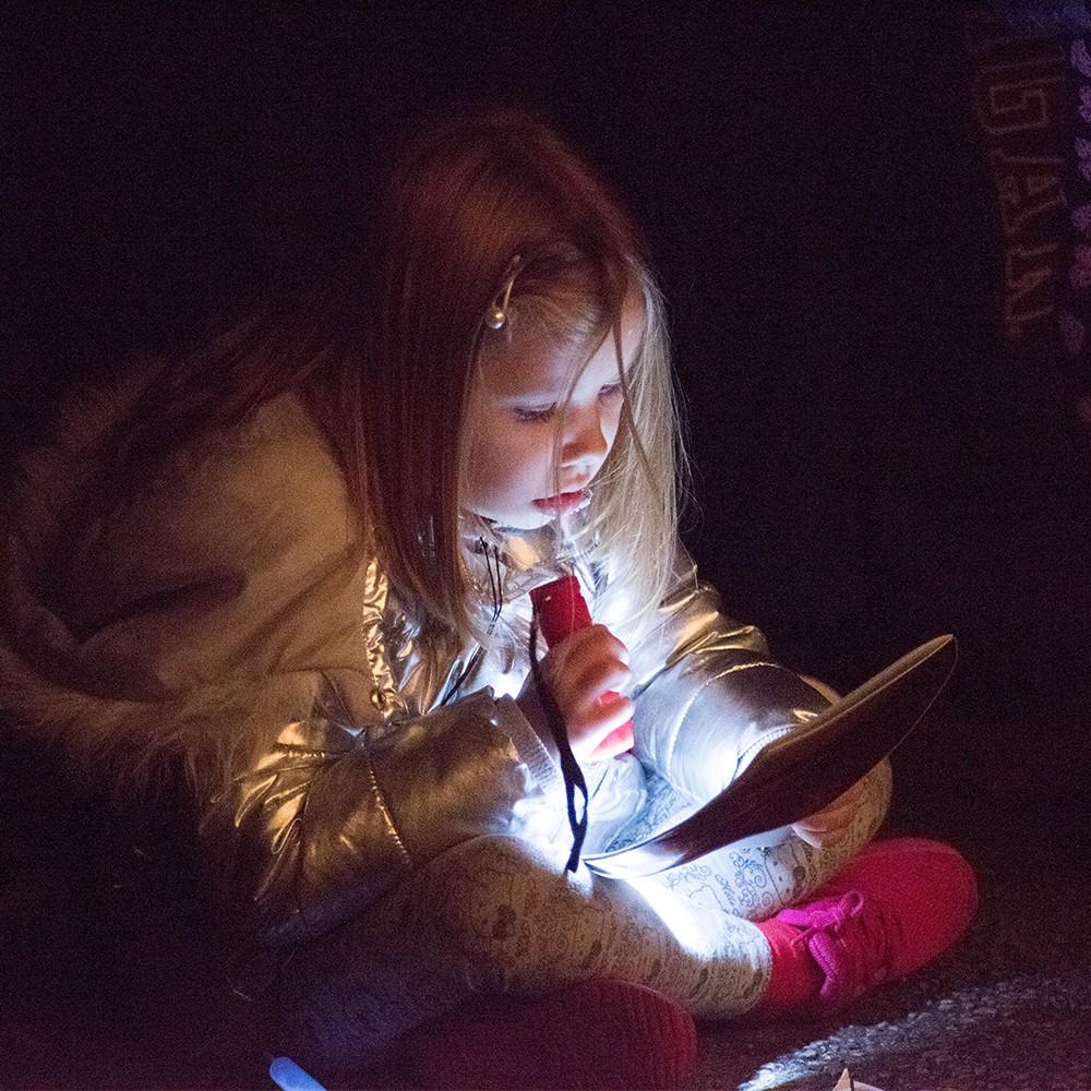 Focused young girl reads a book in the dark, lit by a red flashlight.