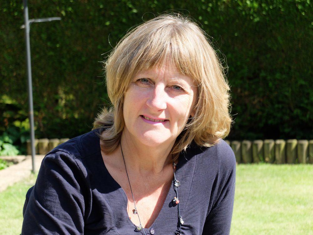 Woman with blonde hair and a dark top smiles in a sunny garden.