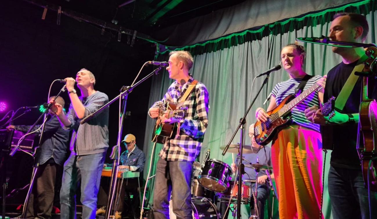 Five musicians perform on stage with guitars, bass, and drums under green and purple lights. Photograph.