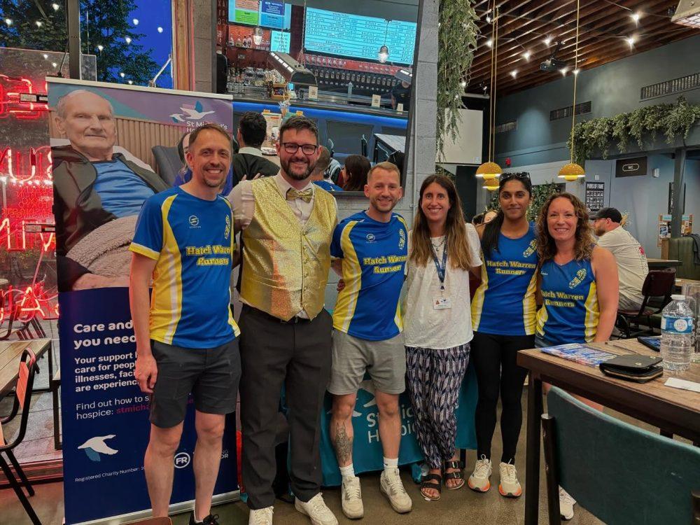 Six smiling people, four in blue and yellow shirts, pose at an indoor event with a banner.