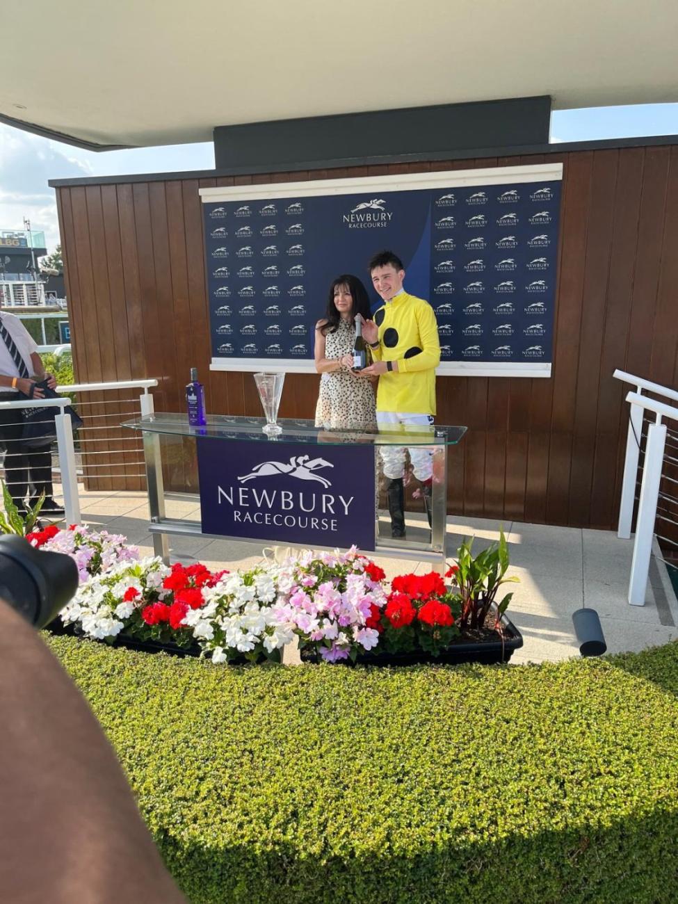 Smiling jockey in yellow silks and woman accept trophies at Newbury Racecourse.