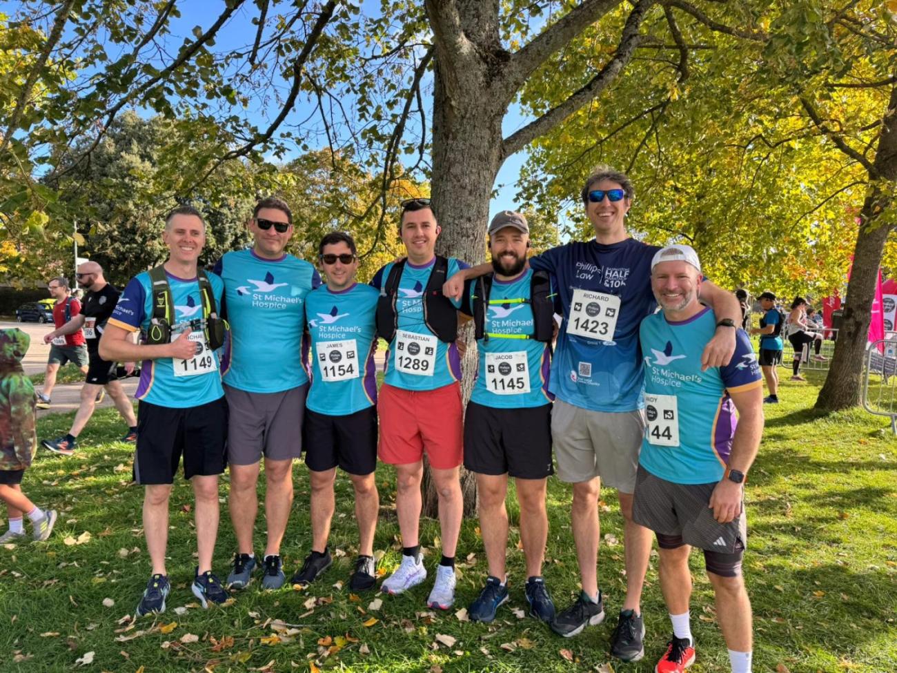 Seven men in matching turquoise shirts with race bibs stand under a tree on a sunny day.