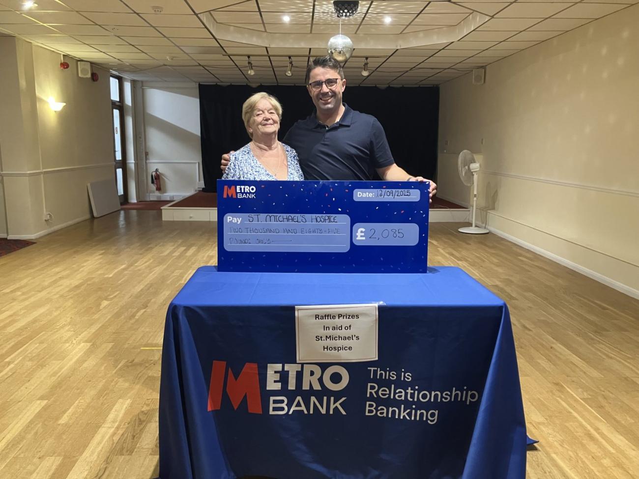 Smiling woman and man behind a large Metro Bank check on a branded table.