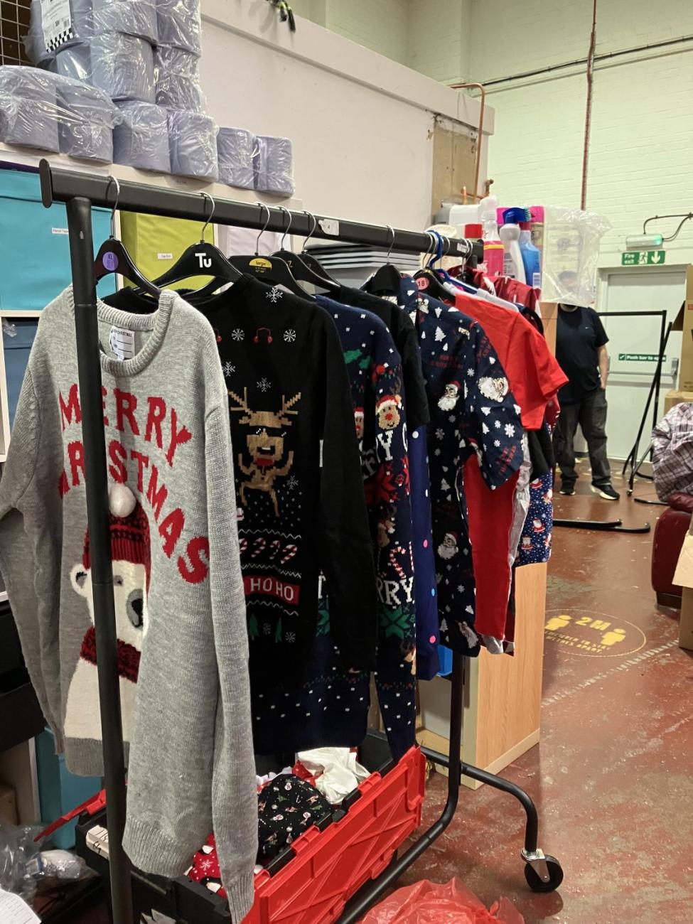 Grey "MERRY CHRISTMAS" sweater and other festive clothes on a rack in a large room.