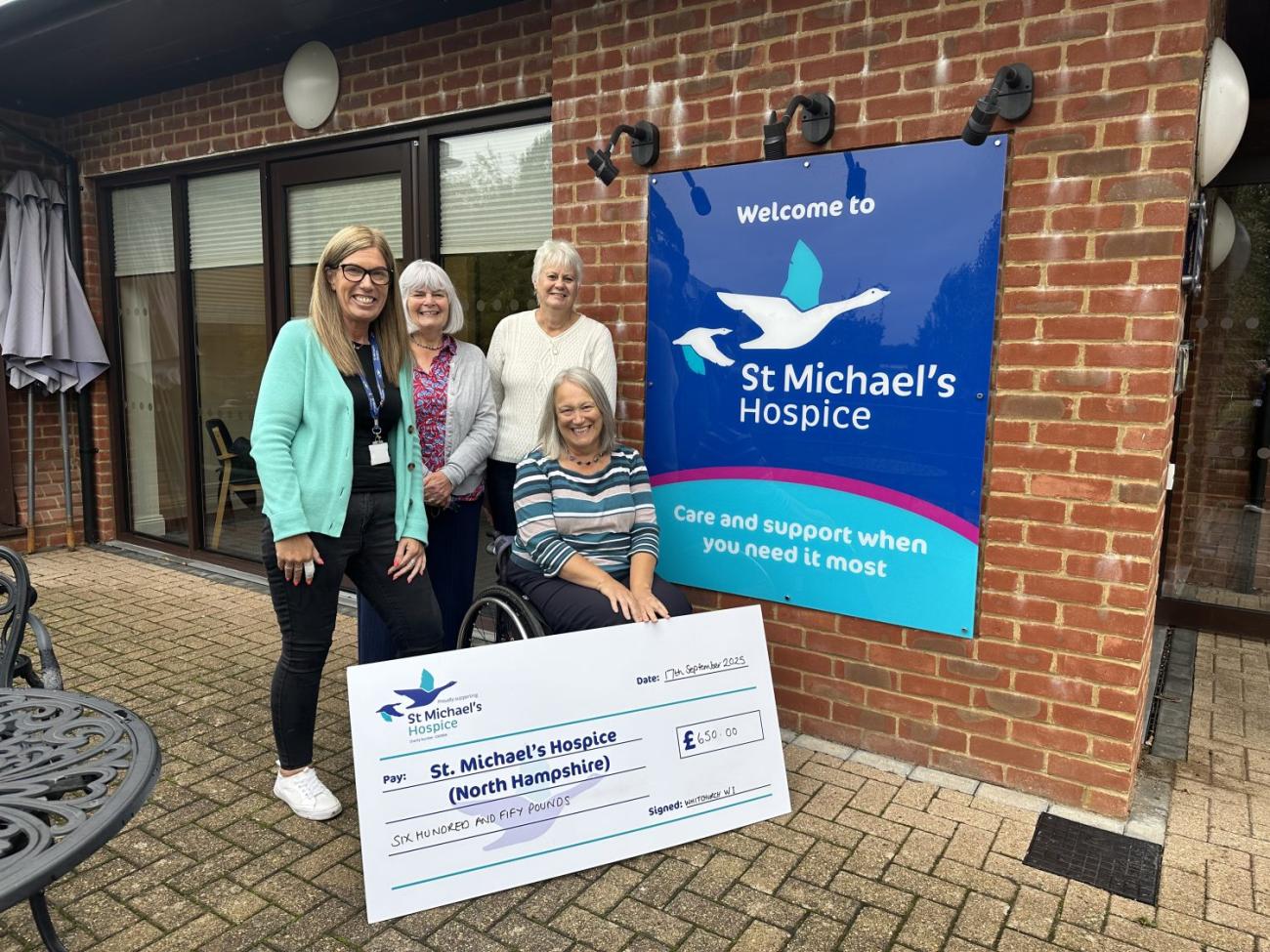 Five women, one in a wheelchair, smiling with a large check for St Michael's Hospice.
