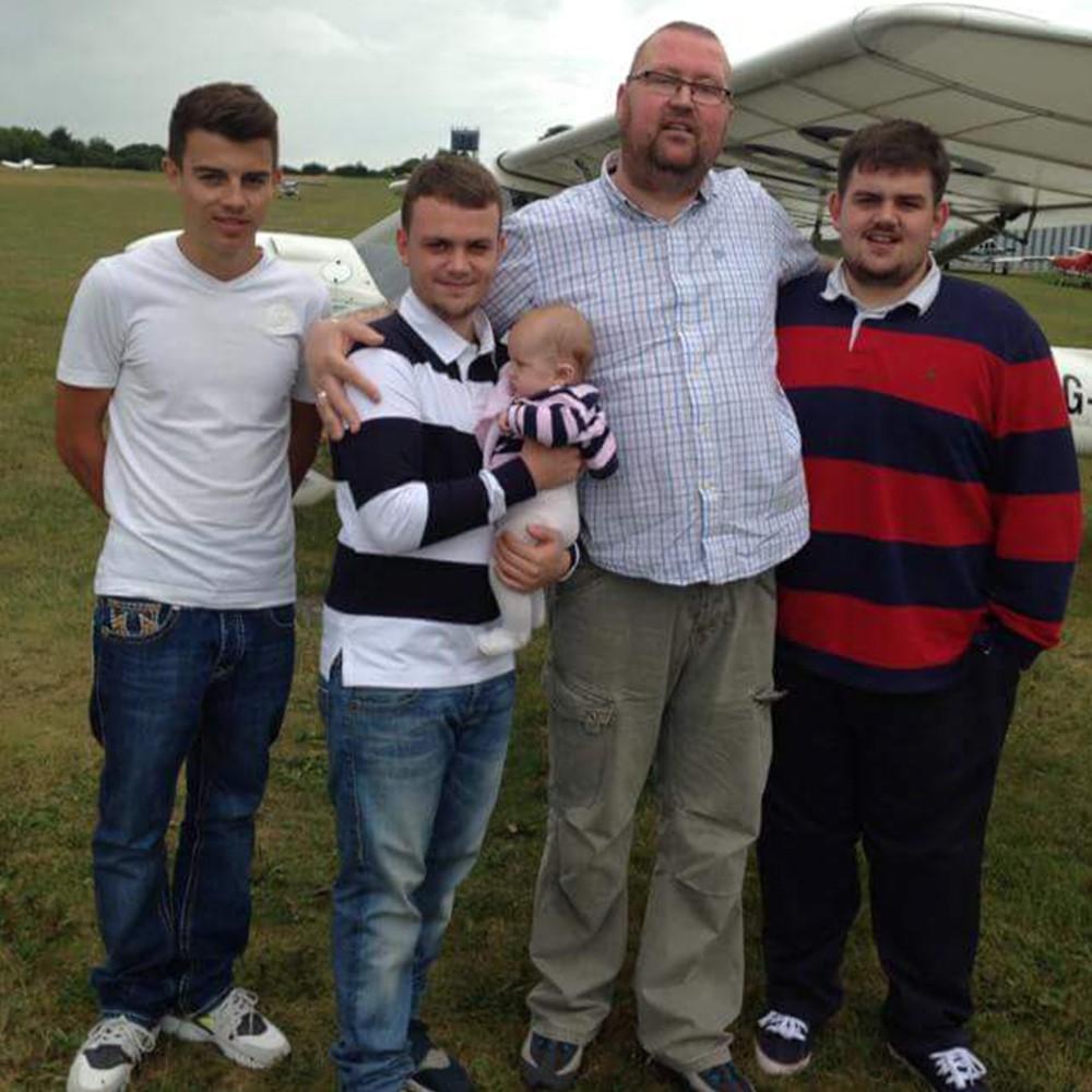 A tall man holding a baby poses with three younger men on a grassy airfield.