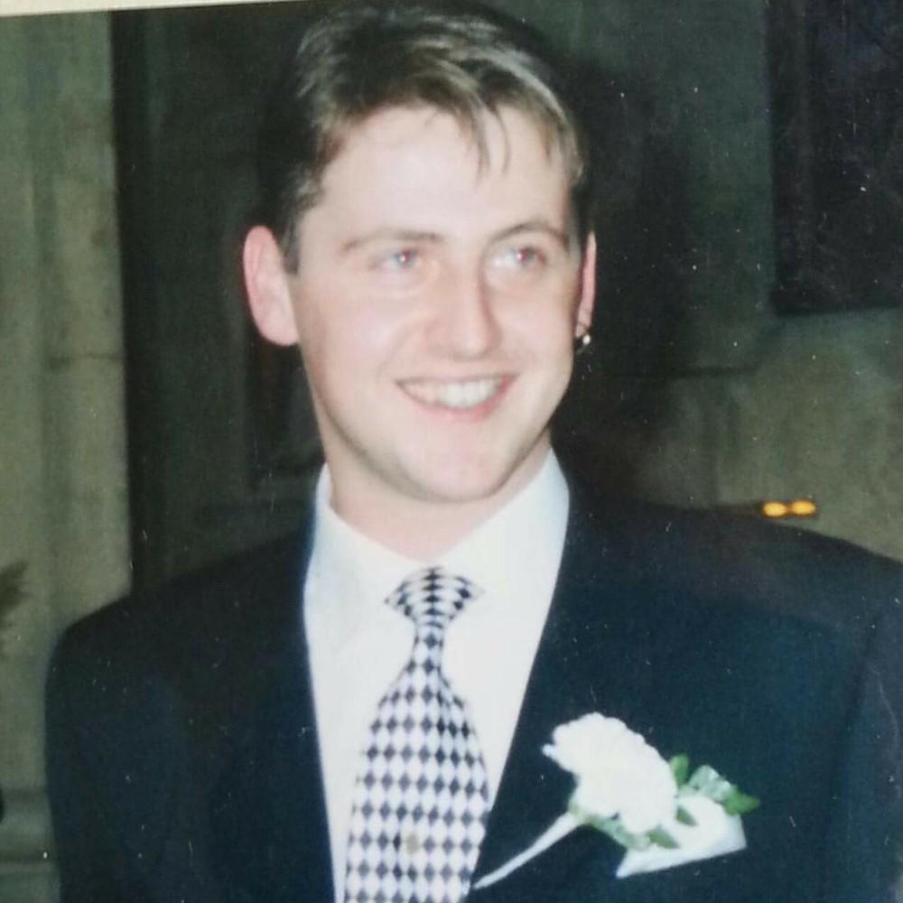 Photograph: Smiling man in a dark suit with a patterned tie and white boutonnière.