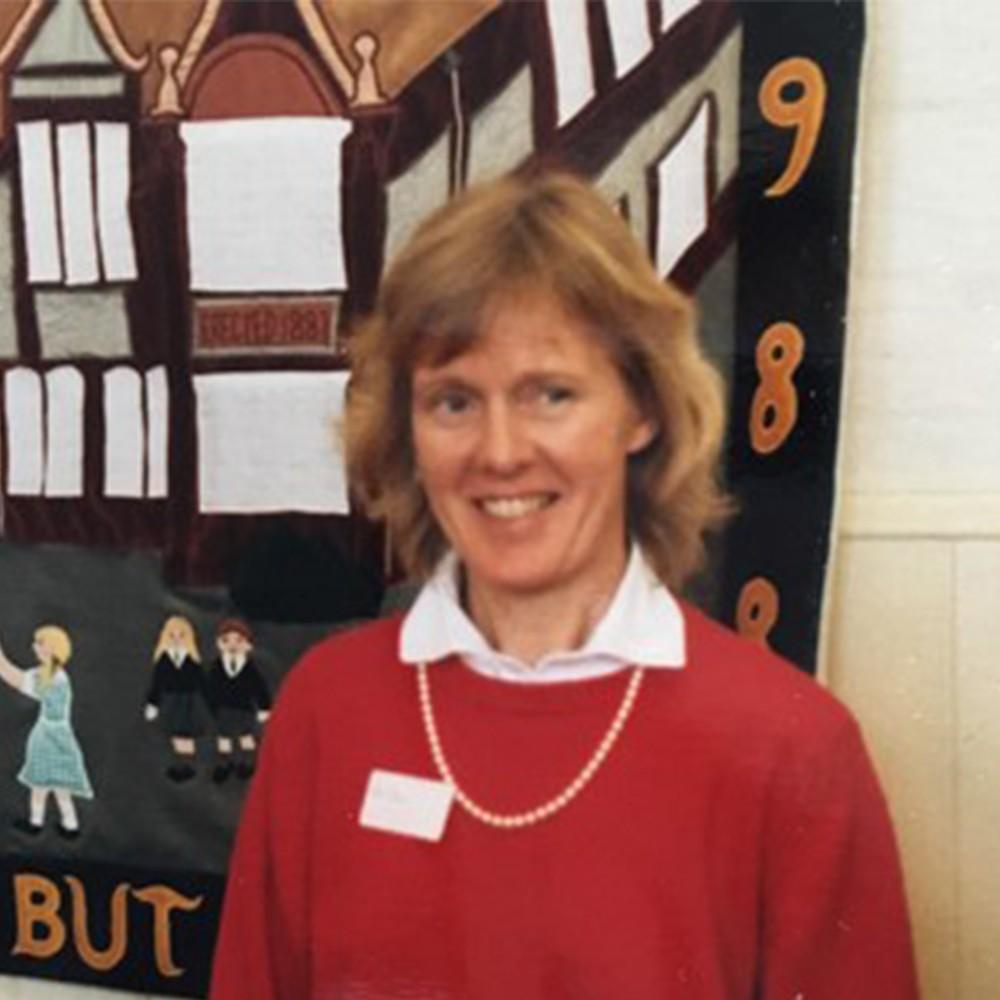 Smiling woman in red sweater and pearl necklace, in front of a textile art piece.