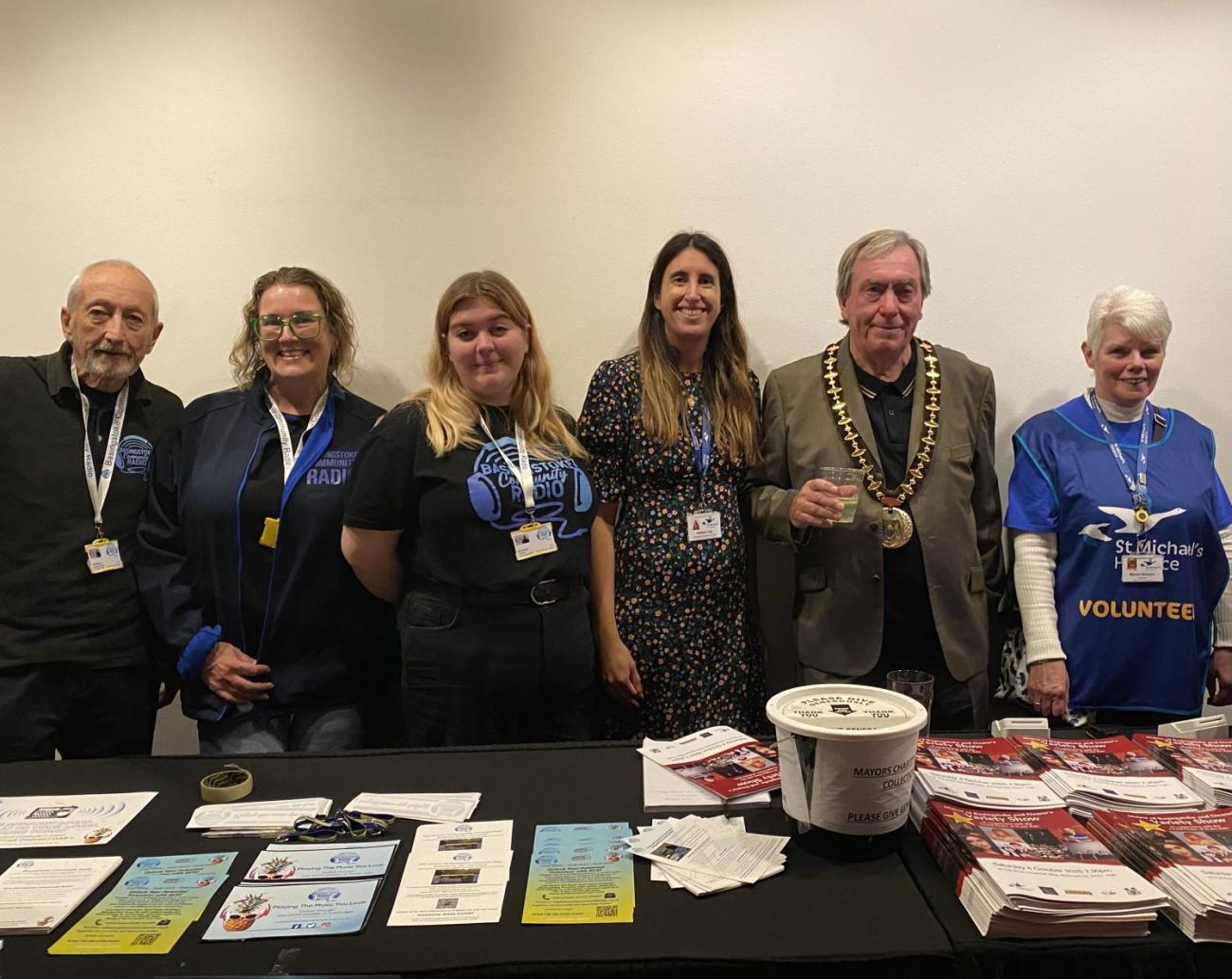 Six people, including a man in a mayoral chain, stand behind an event table with brochures.