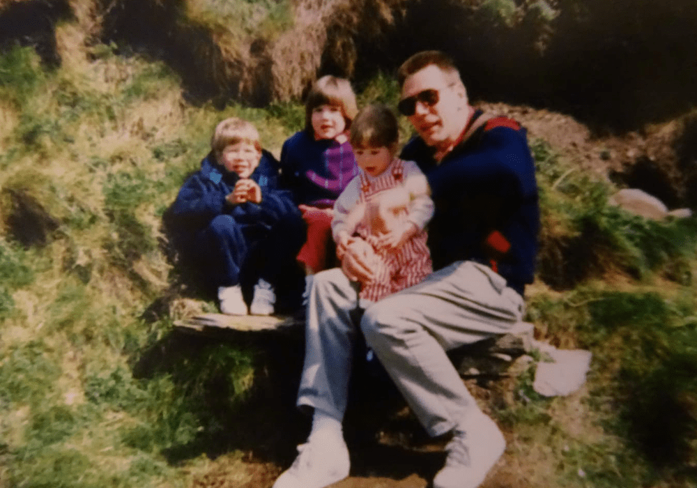 Dad holds a toddler with two other children nearby on a sunny, grassy hillside.