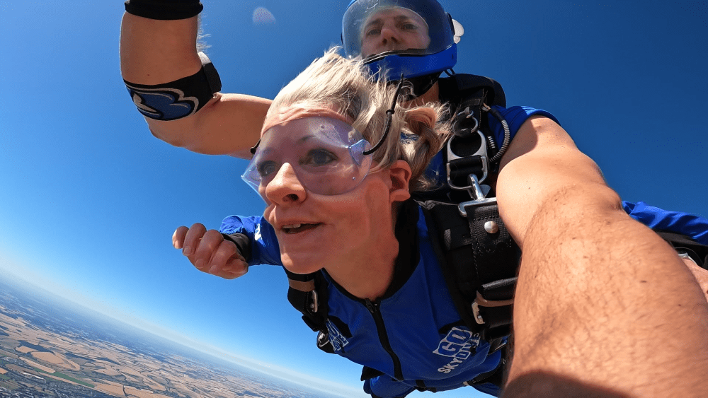 Smiling woman and instructor skydiving in blue suits against a clear blue sky.
