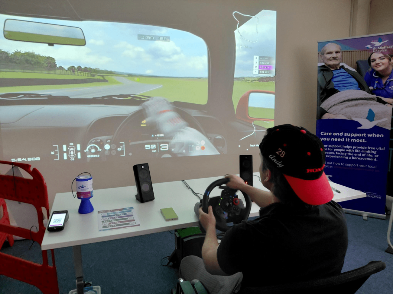 Person in a cap plays a driving simulator, game projected onto a large wall.
