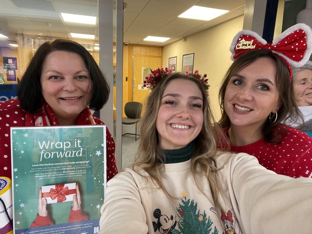Selfie of three smiling women in festive attire, one holding a poster with a gift.