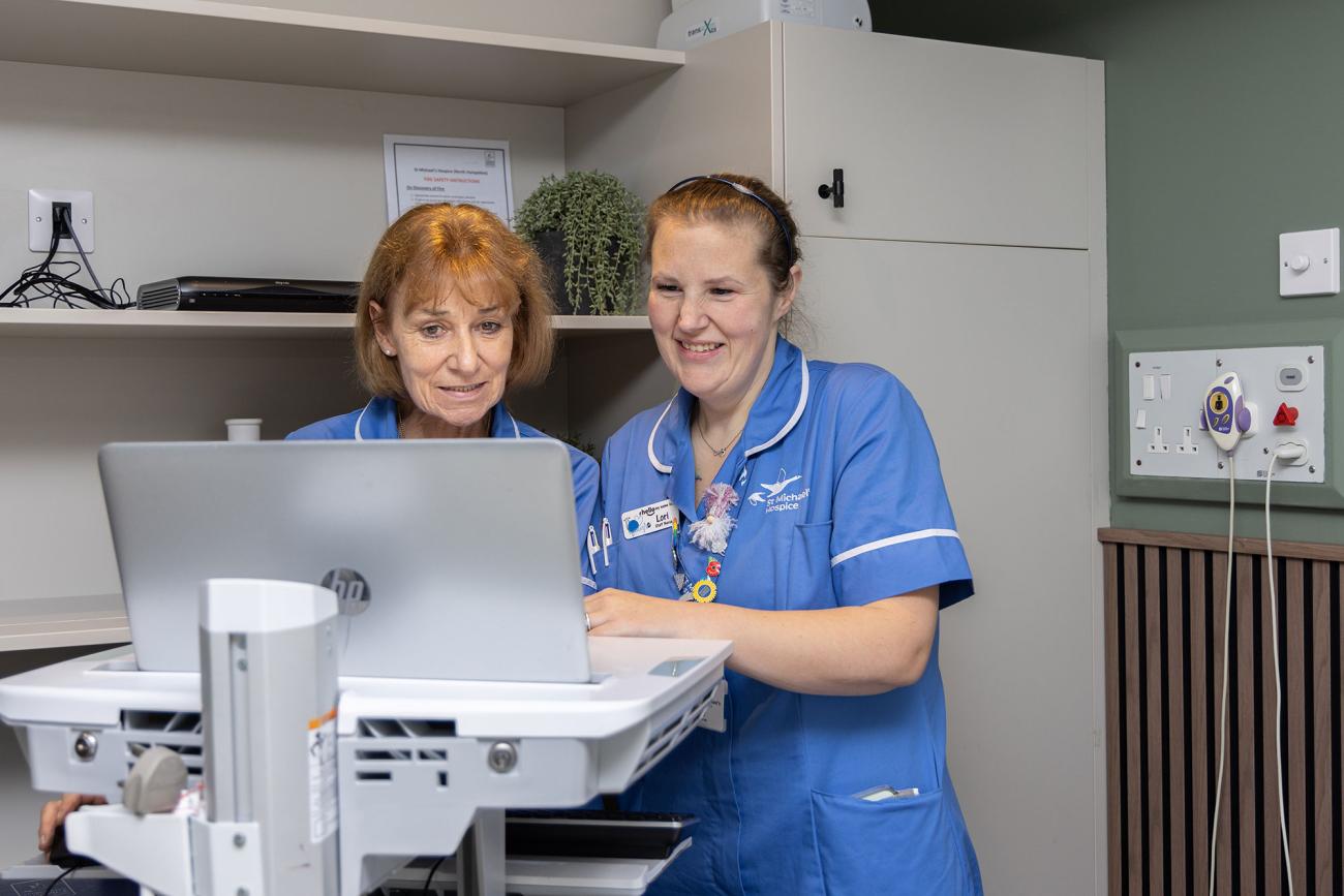 Two smiling nurses in blue scrubs look at a laptop on a medical cart in a clinic.