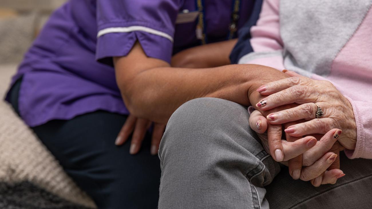 A person in a purple uniform gently holds the hand of an elderly person.