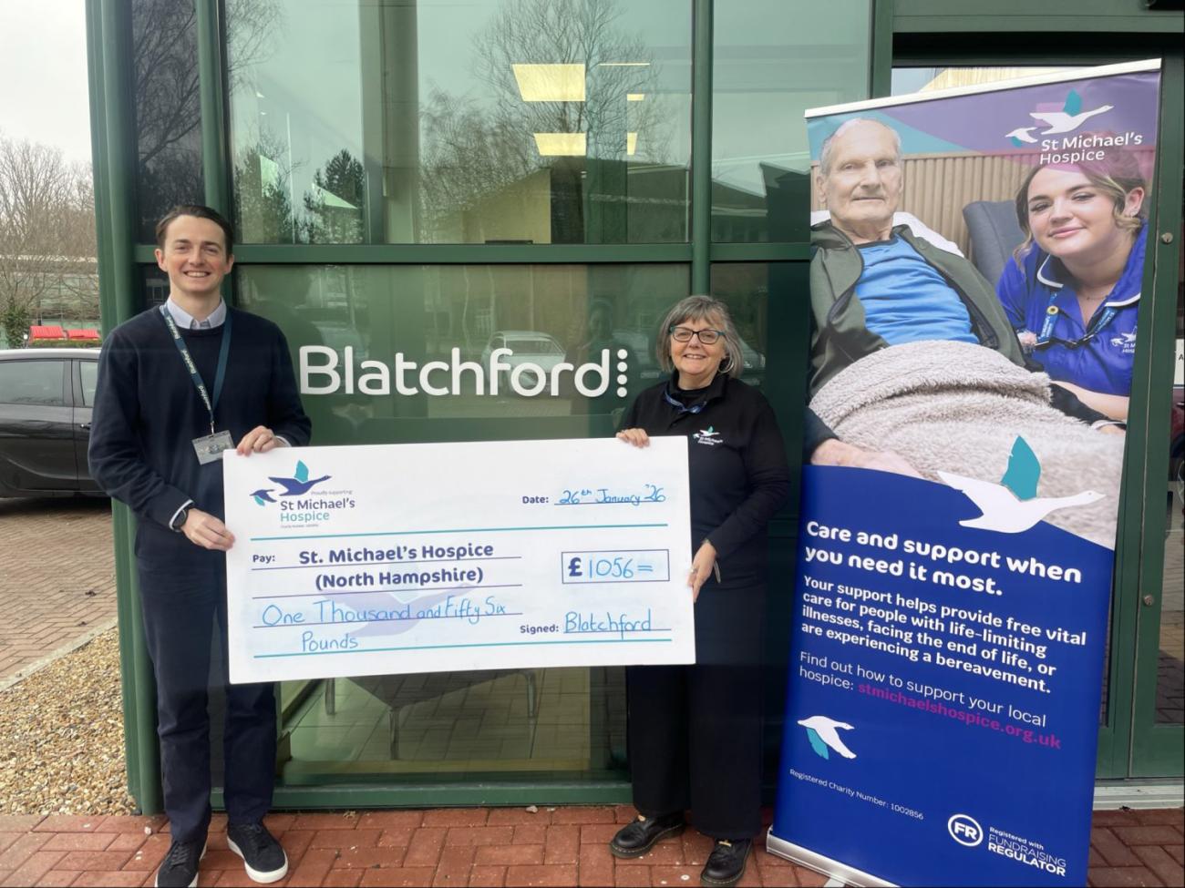 Man and woman hold a large check for St. Michael's Hospice outside Blatchford building.