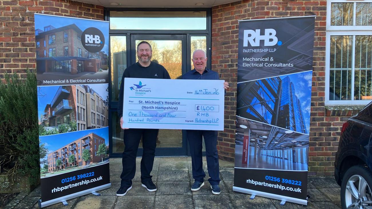 Two smiling men hold a large check for £1,000, flanked by RHB Partnership banners.