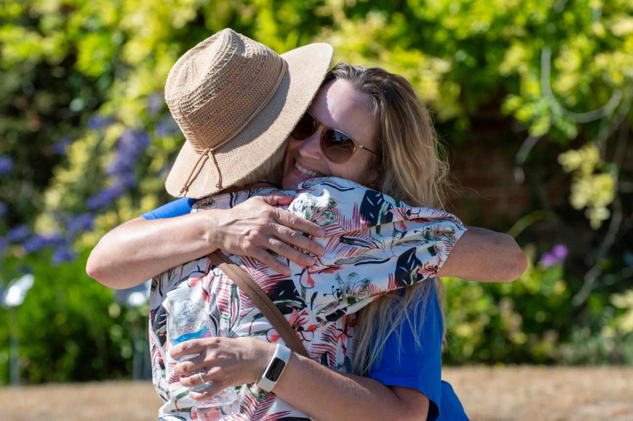 Two people sharing a warm hug outdoors, one in a hat, the other in sunglasses.