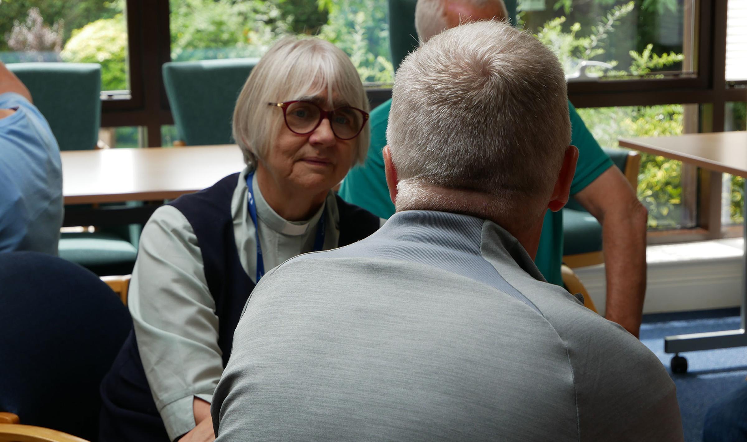Woman with glasses intently listening to a man at an indoor gathering.